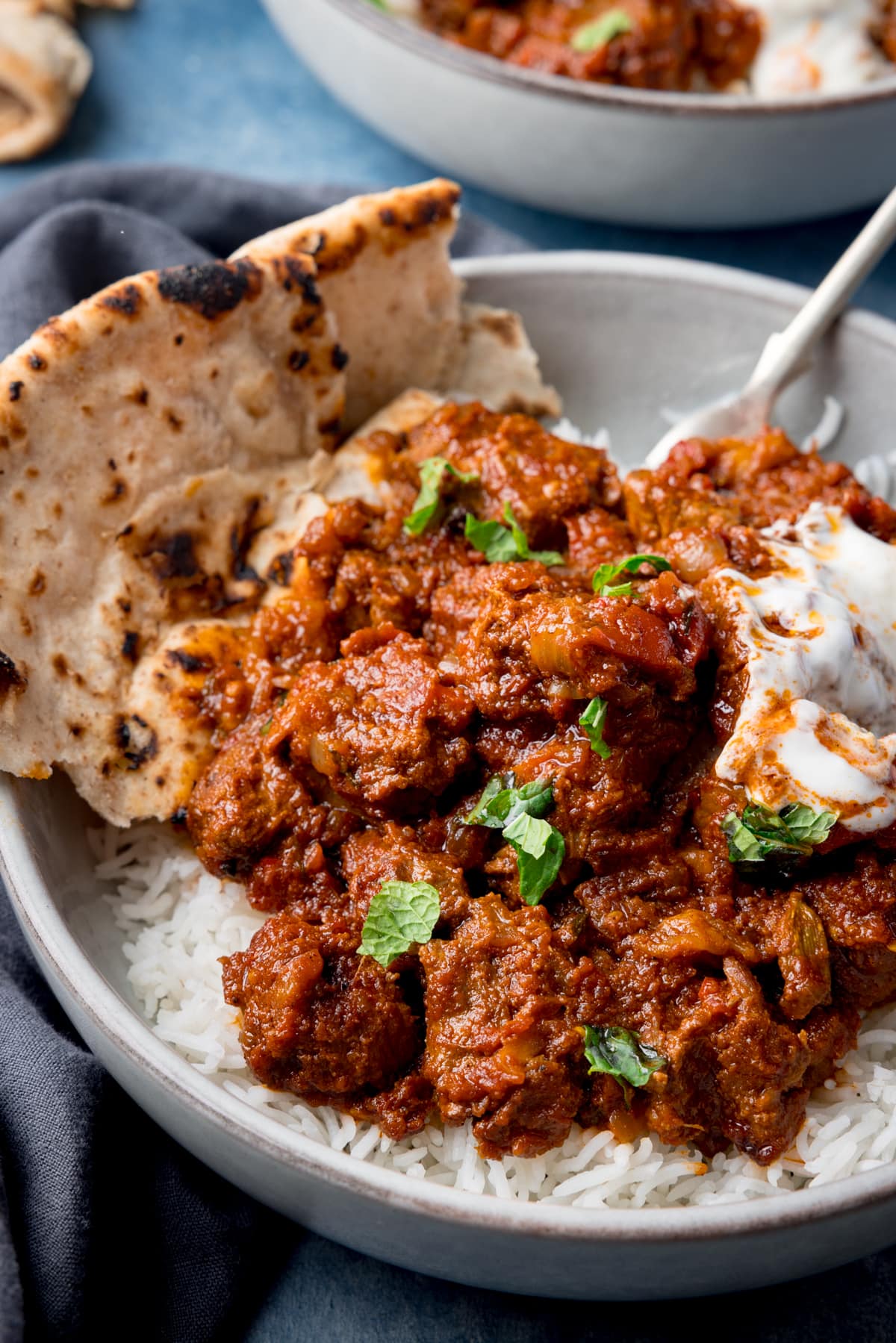 Tall image of beef rogan josh and rice in a white bowl with a piece of chapati on the side. The bowl is on a blue background, next to a blue/grey napkin. There is a further bowl at the top of the image.