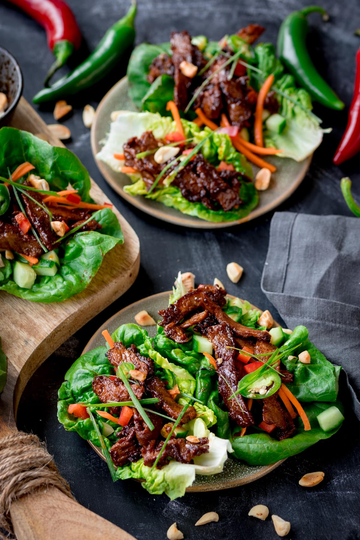 Picture of some Korean beef lettuce wraps on a small green plate with another plate and some chilli's in the background. All sat on a blue board.