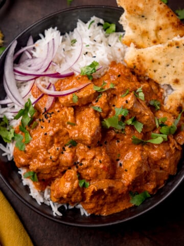 Overhead picture of a dark bowl full of boiled rice and crock pot butter chicken with some sliced red onion and charred naan bread in the corner of the bowl.