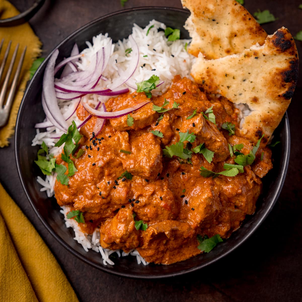 Overhead picture of a dark bowl full of boiled rice and crock pot butter chicken with some sliced red onion and charred naan bread in the corner of the bowl.
