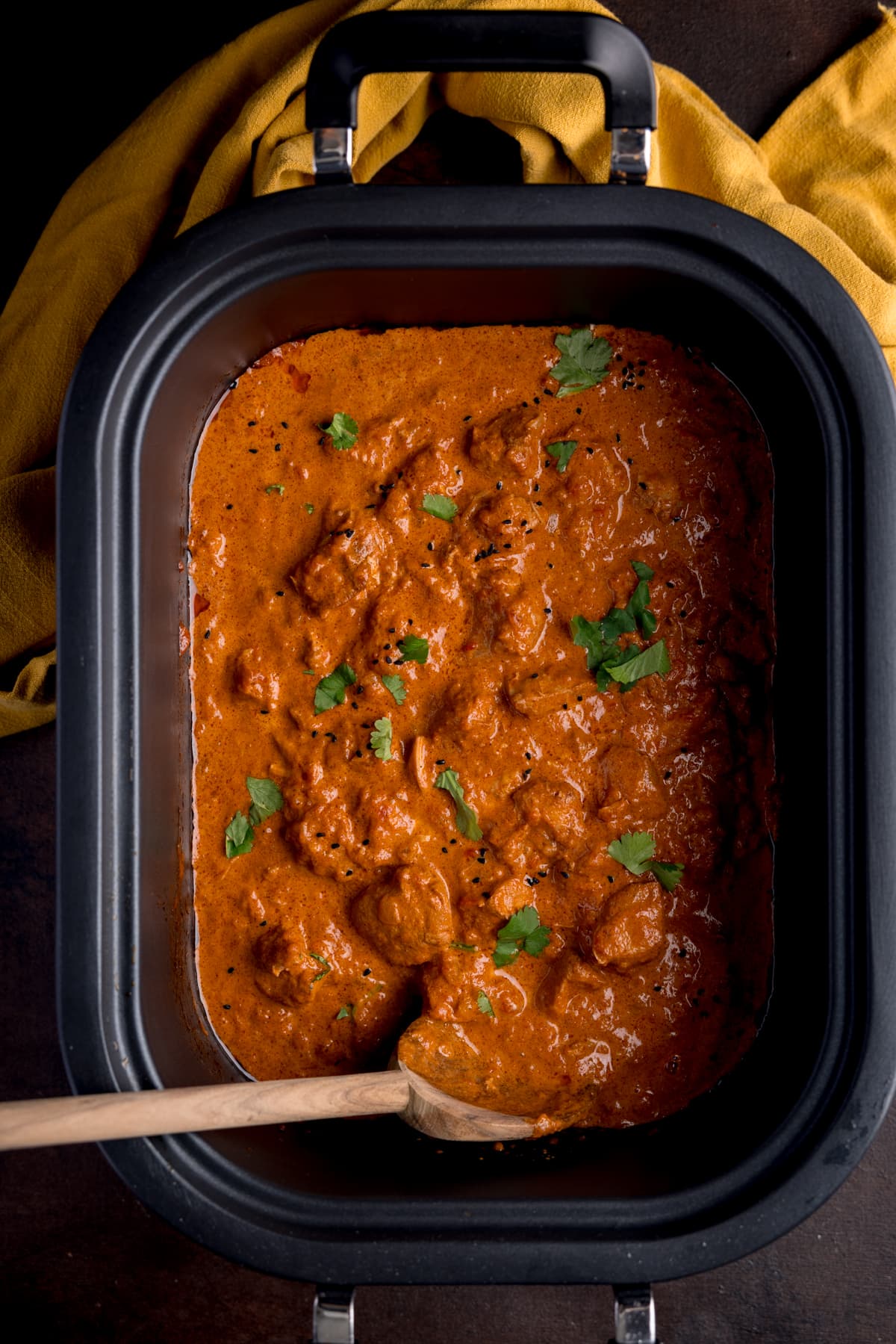 Overhead picture of crock pot butter chicken in the slow cooker bowl, sprinkled with a little fresh coriander with a wooden spoon in the corner.