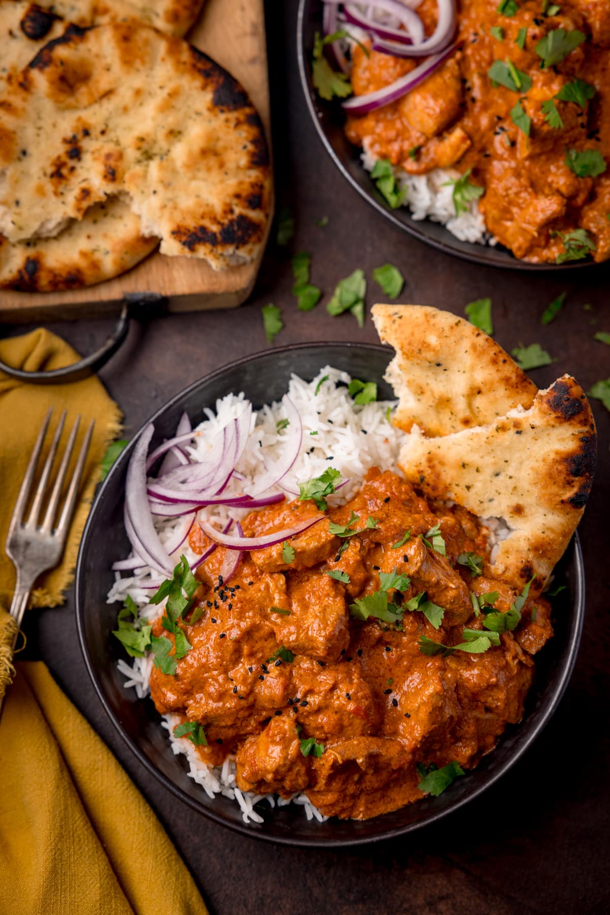 Overhead picture of a dark bowl full of boiled rice and crock pot butter chicken with some sliced red onion and charred naan bread in the corner of the bowl, with a fork and a board with more naan bread in the background.