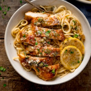 Square image of lemon chicken piccata on a bed of linguine with slices of lemon in a white bowl. The chicken has been sliced into strips. The bowl is on a wooden table.