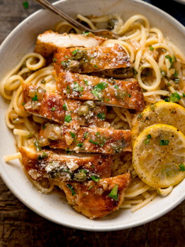 Square image of lemon chicken piccata on a bed of linguine with slices of lemon in a white bowl. The chicken has been sliced into strips. The bowl is on a wooden table.