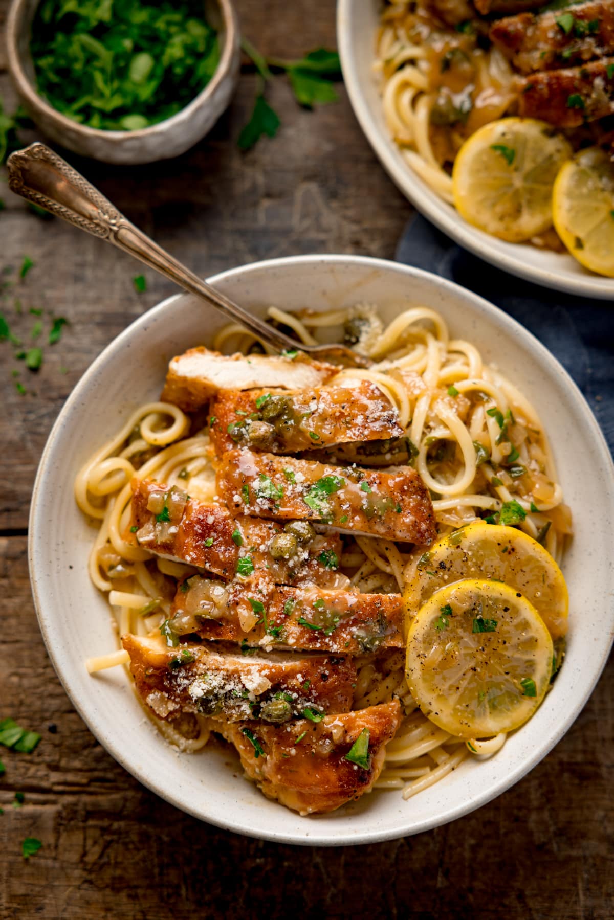 Tall overhead image of lemon chicken piccata on a bed of linguine with slices of lemon in a white bowl. The chicken has been sliced into strips and there is a fork sticking out. The bowl is on a wooden table next to a small bowl of herbs. There is a further bowl of lemon chicken piccata just in shot at the top of the frame.