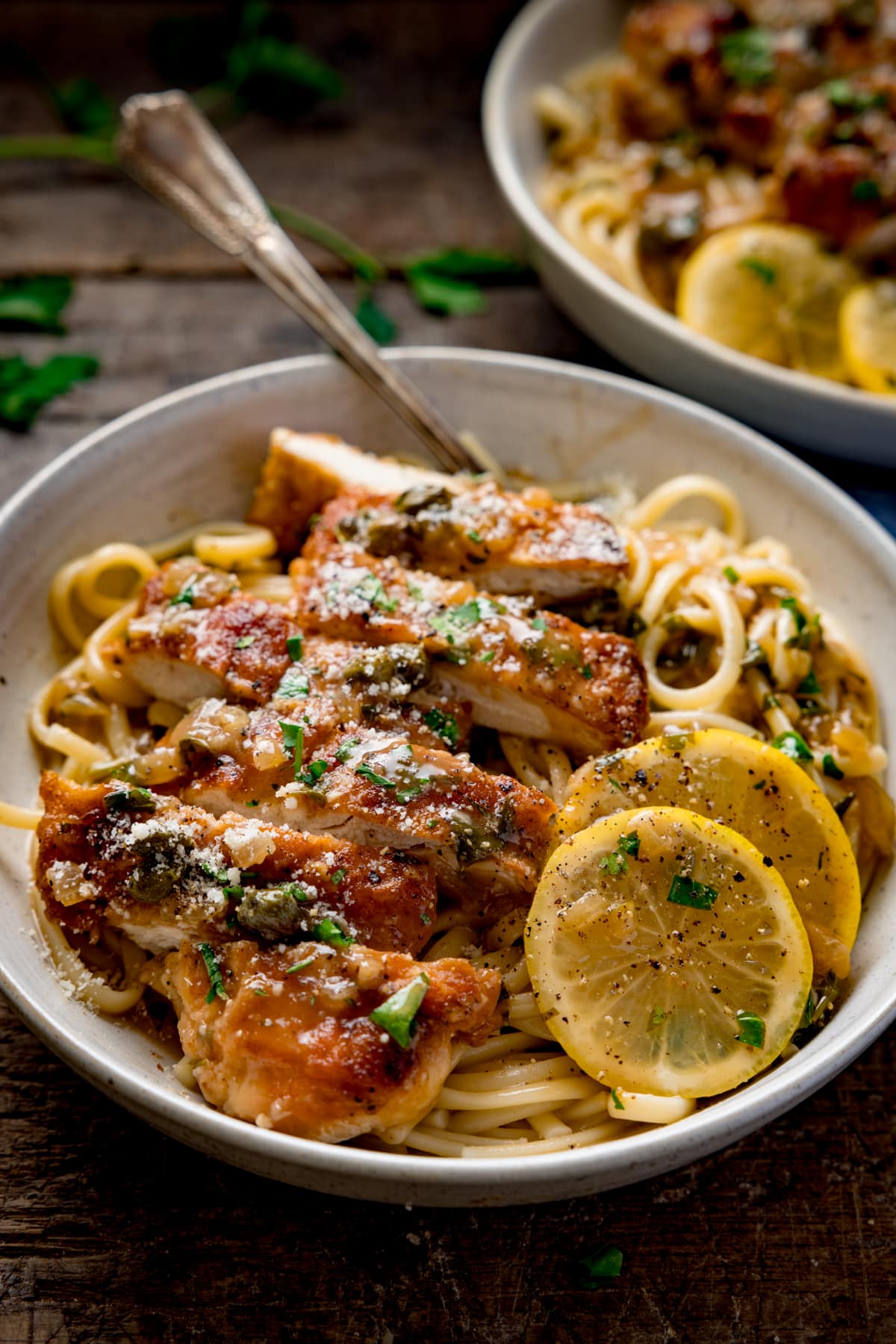 Tall image of lemon chicken piccata on a bed of linguine with slices of lemon in a white bowl. The chicken has been sliced into strips. The bowl is on a wooden table and there is a fork sticking out. There is a second bowl in the background.