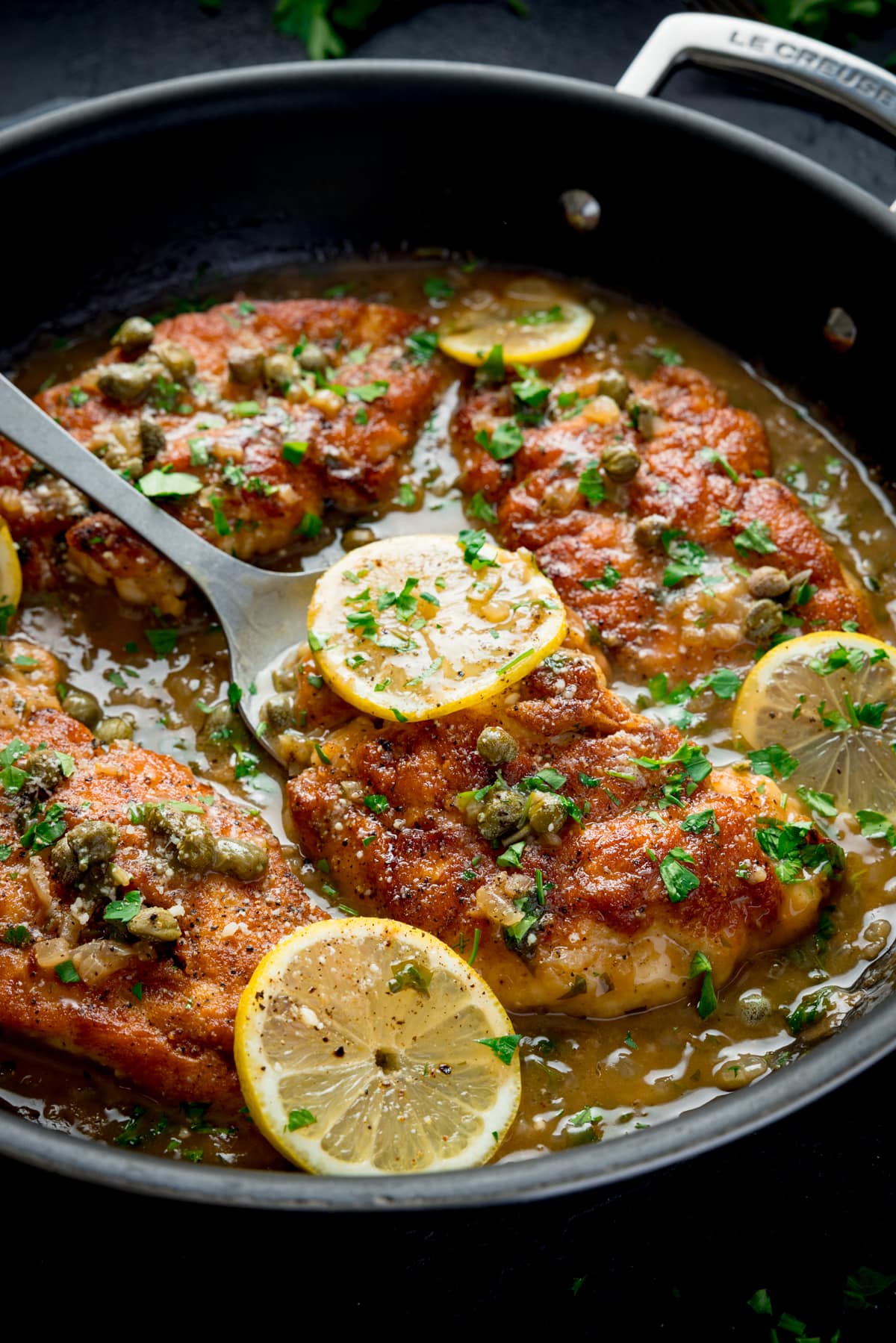Lemon chicken piccata in a black pan topped with lemon sliced and parsley. There is a spoon starting to lift up one of the pieces of chicken.