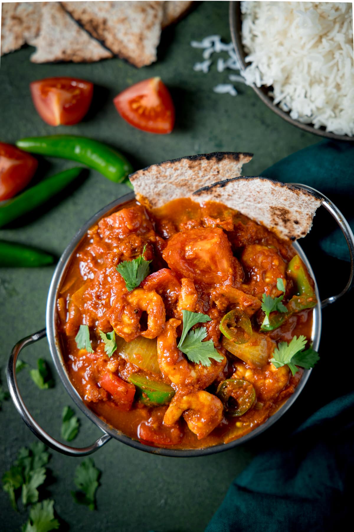 Overhead picture of a prawn balti in a silver balti dish on a green background with some rice and toasted chapati in the background.