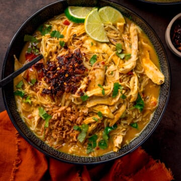 Overhead picture of a black textured bowl full of chicken laksa topped with some chilli crisp and a sprinkling of fresh coriander and some lime wedges.