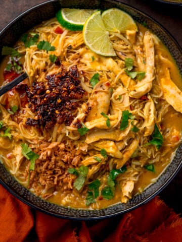Overhead picture of a black textured bowl full of chicken laksa topped with some chilli crisp and a sprinkling of fresh coriander and some lime wedges.