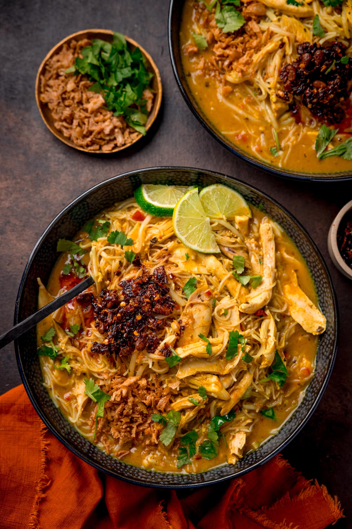 Overhead picture of a black textured bowl full of chicken laksa topped with some chilli crisp and a sprinkling of fresh coriander and some lime wedges and an orange linen cloth in the foreground.