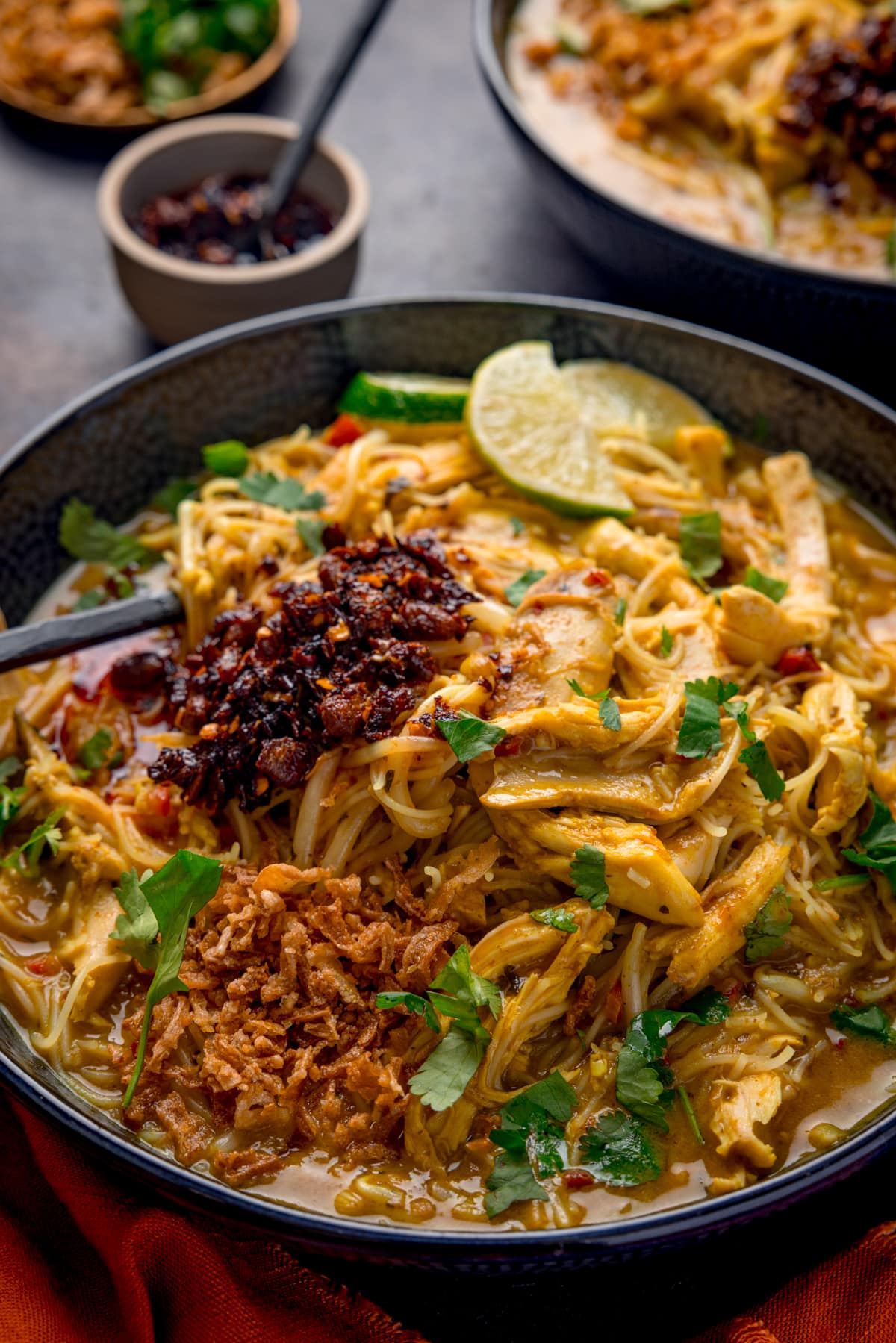 Close up picture of some quick chicken laksa in a dark bowl, topped with crispy onions, chilli crisp and fresh coriander.
