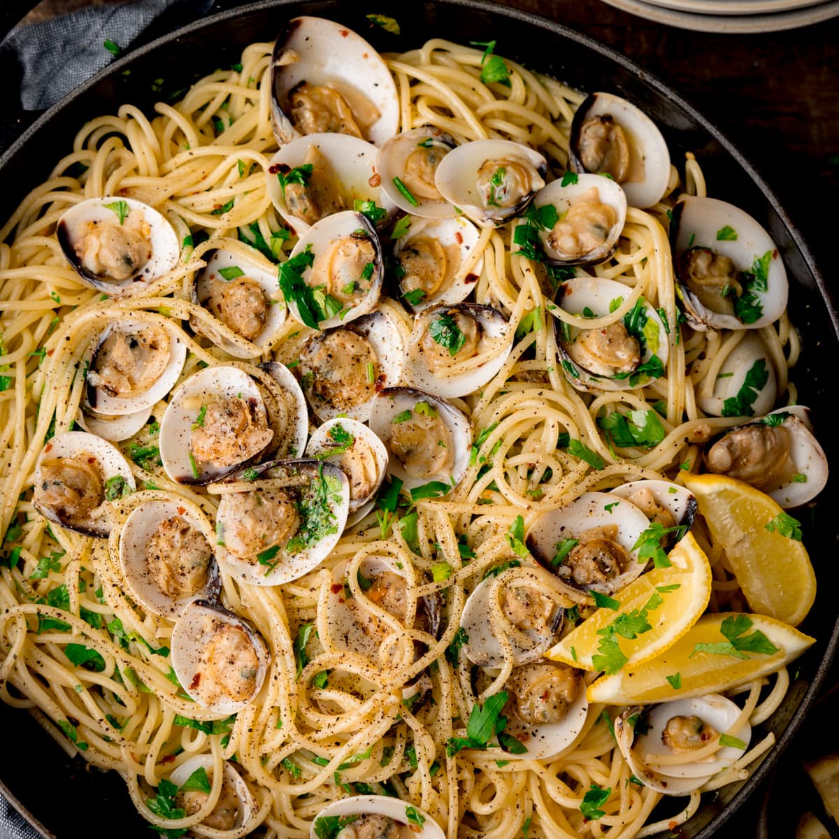 Overhead square image of Spaghetti Vongole (spaghetti with clams) in a black pan topped with parsley and lemon wedges.