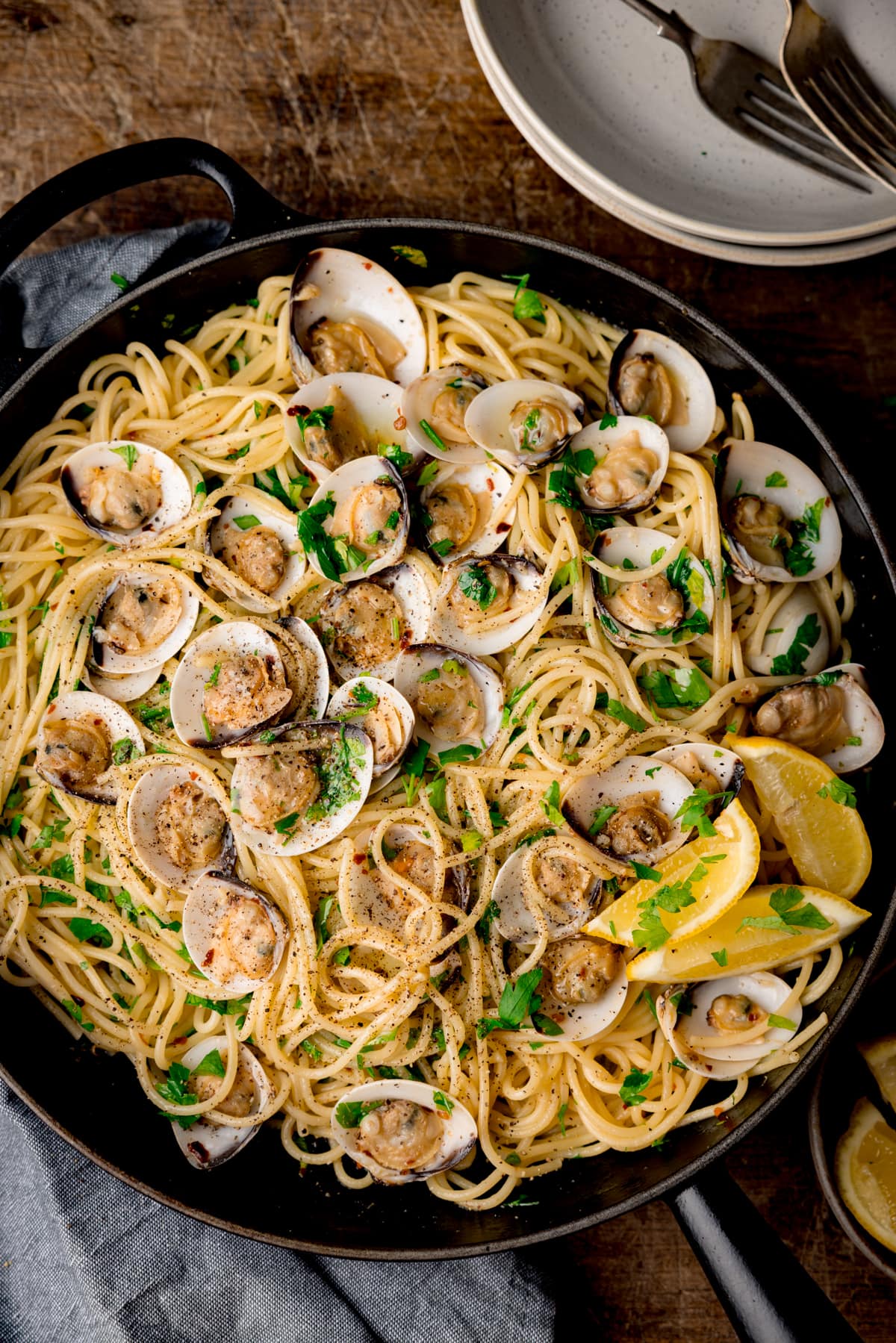 Tall overhead image of Spaghetti Vongole (spaghetti with clams) in a black pan topped with parsley and lemon wedges. The pan is on a wooden table next to a grey napkin. There are two white bowls with forks at the top of the frame.