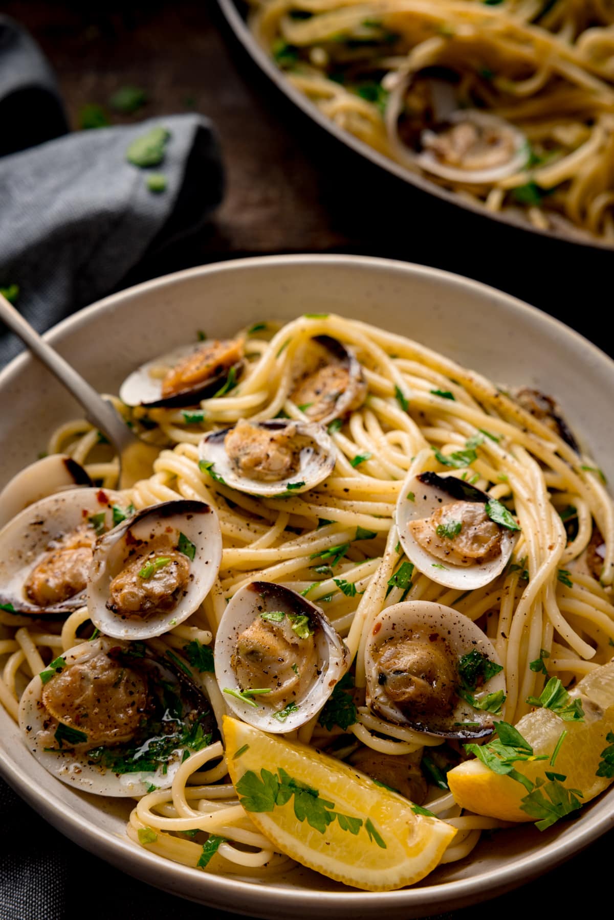 Spaghetti vongole (spaghetti with clams) in a white bowl with a fork sticking out. The spaghetti is topped with parsley and there are lemon wedges in the bowl. The bowl is on a wooden table next to a grey napkin. The pan of spaghetti vongole is just in shot at the top of the frame.