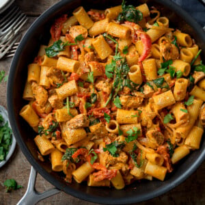 Square overhead image of Tuscan Chicken Pasta in a black pan on a wooden background. There is parsley and forks scattered around the pan.