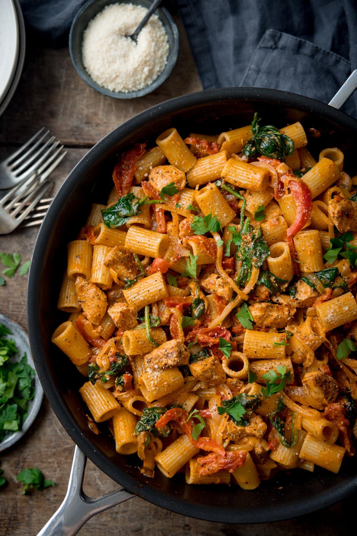 Tall overhead image of Tuscan Chicken Pasta in a black pan on a wooden background. There is parsley and forks scattered around the pan. The pan is next to a blue napkin and there is a bowl of grated parmesan with a spoon sticking out at the top of the frame.