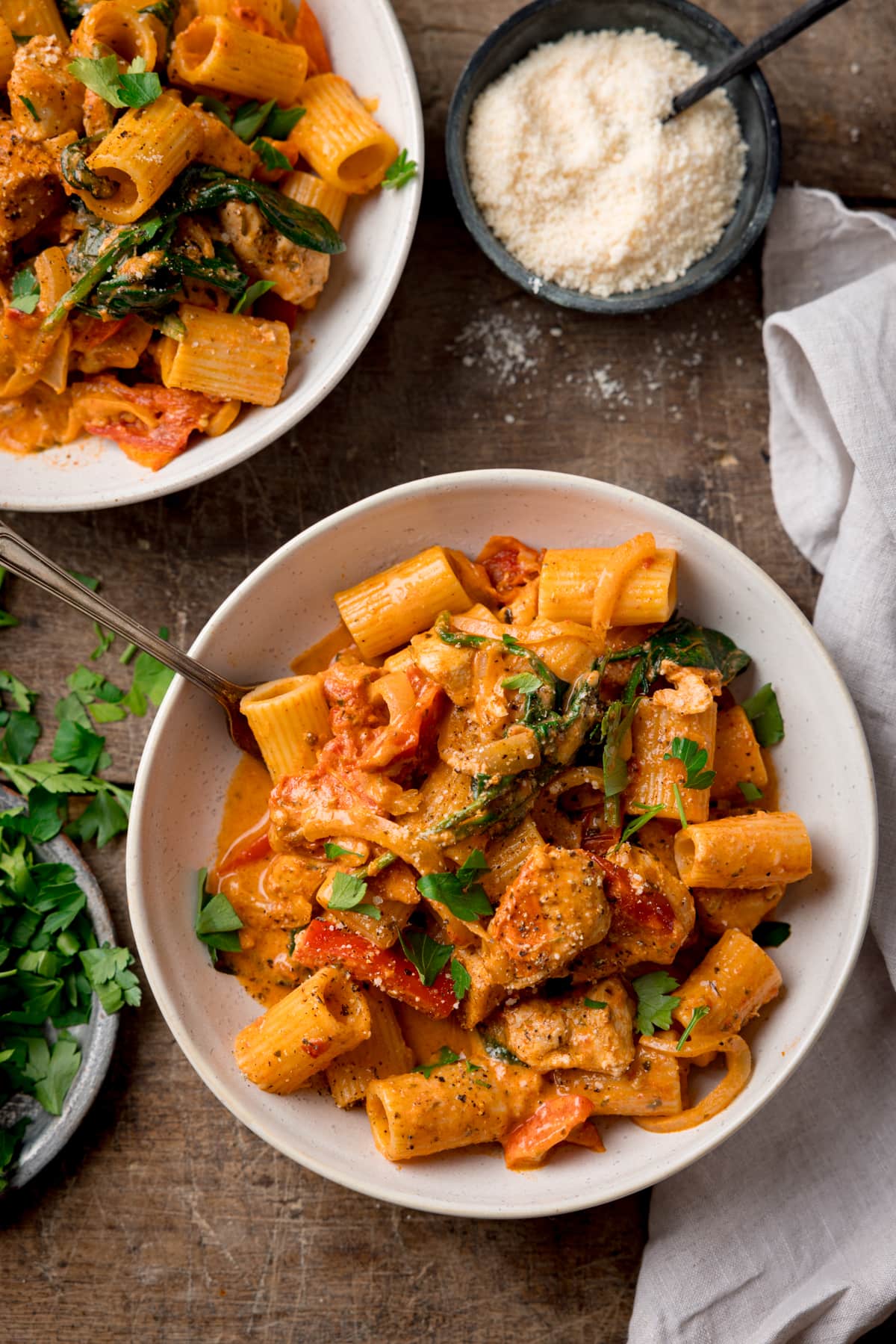 A white bowl filled with Tuscan Chicken Pasta on a wooden background. There us a fork sticking out of the dish. There is a further bowl of pasta at the top of the frame, next to a bowl of grated parmesan. There is a also a light napkin to the right of the main bowl and a small silver plate of chopped parsley on the left of the main bowl.