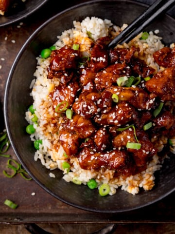 Square image of air fryer sesame chicken served on a bed of egg fried rice in a black bowl. The sesame chicken is topped with sesame seeds and spring onions. There is a pair of black chopsticks sticking out of the bowl. The bowl is on a dark background. There is a small plate with spring onions on the the left of the frame.