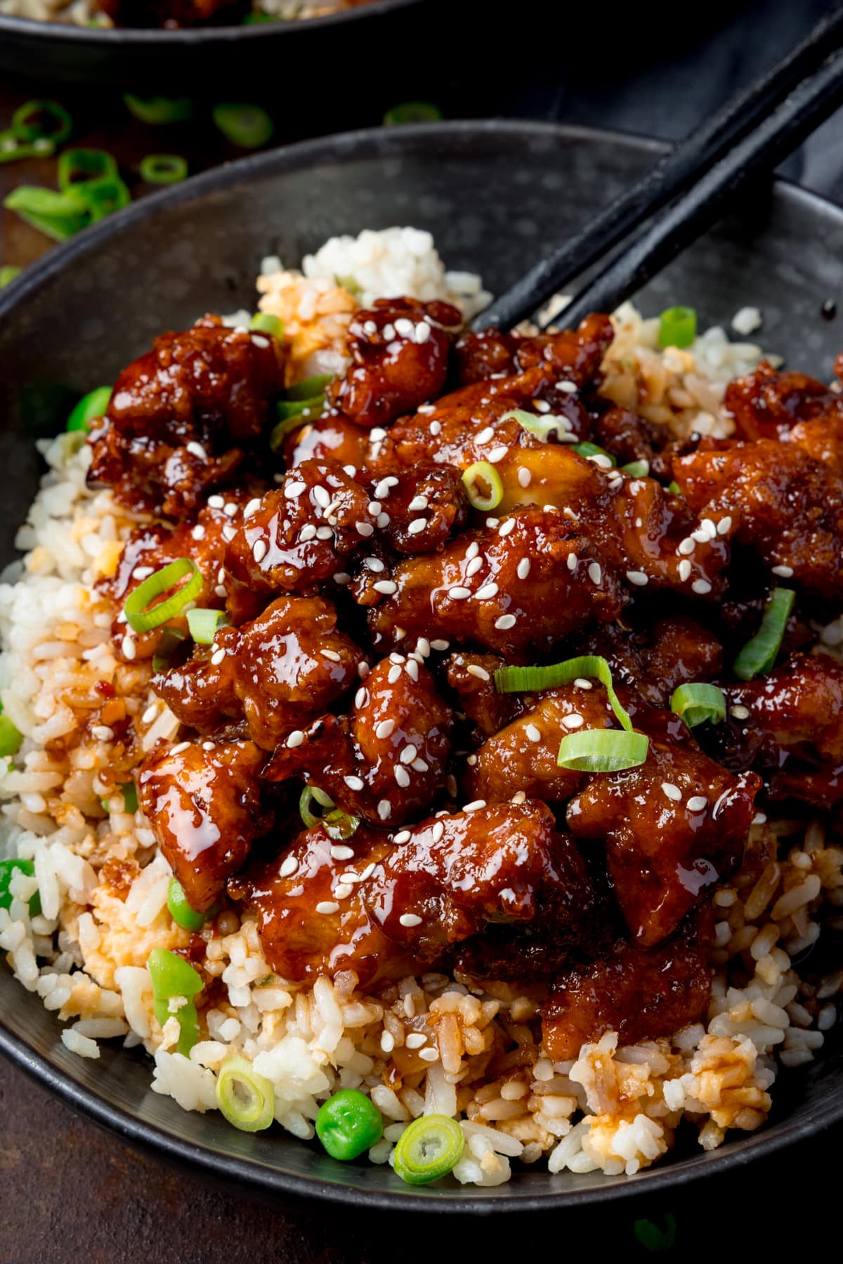 Tall close-up image of air fryer sesame chicken served on a bed of egg fried rice in a black bowl. The sesame chicken is topped with sesame seeds and spring onions. There is a pair of black chopsticks sticking out of the bowl. The bowl is on a dark background.