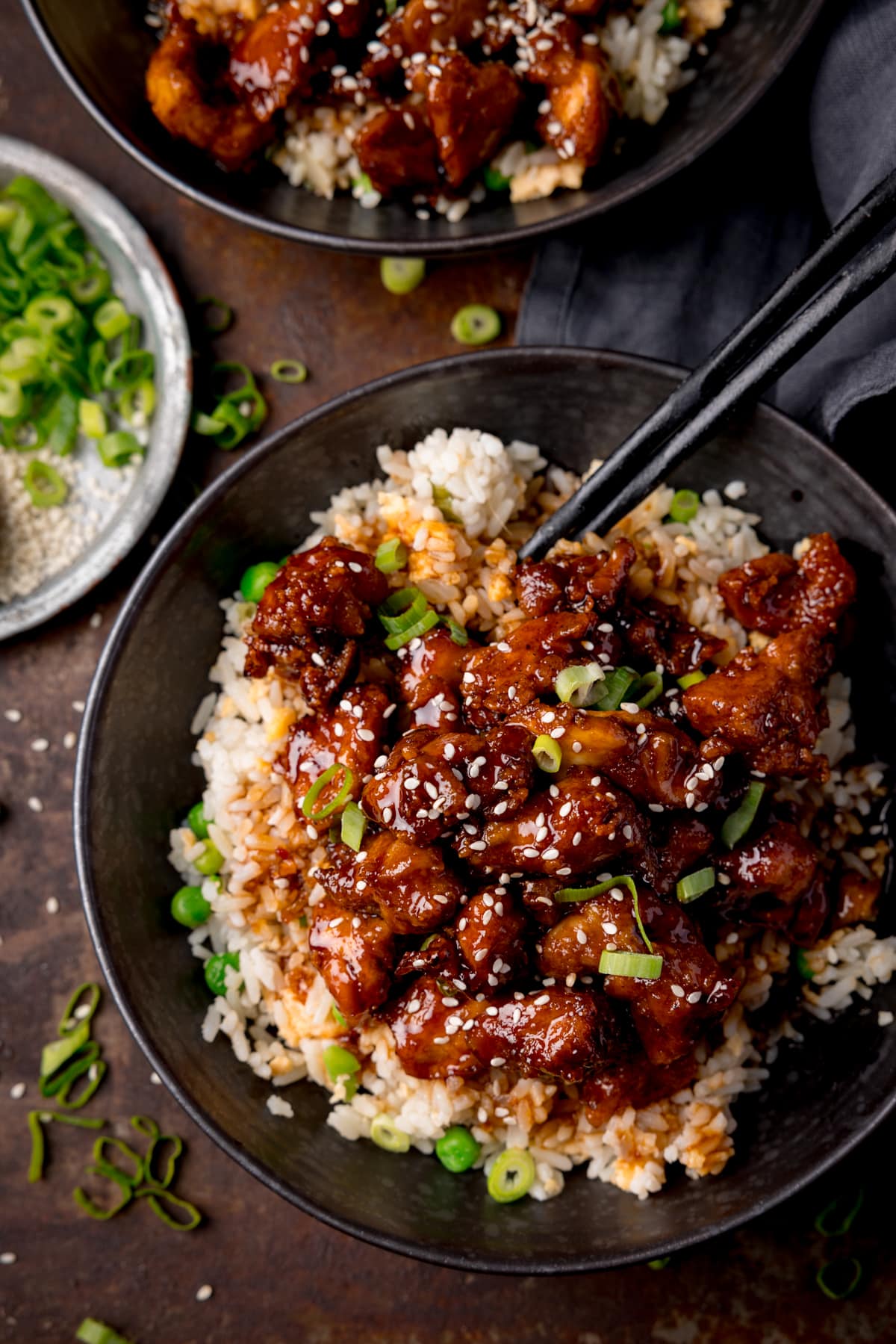 Tall overhead image of air fryer sesame chicken served on a bed of egg fried rice in a black bowl. The sesame chicken is topped with sesame seeds and spring onions. There is a pair of black chopsticks sticking out of the bowl. The bowl is on a dark background. There is a further bowl of sesame chicken at the top of the frame and a small plate with spring onions on the the left of the frame.