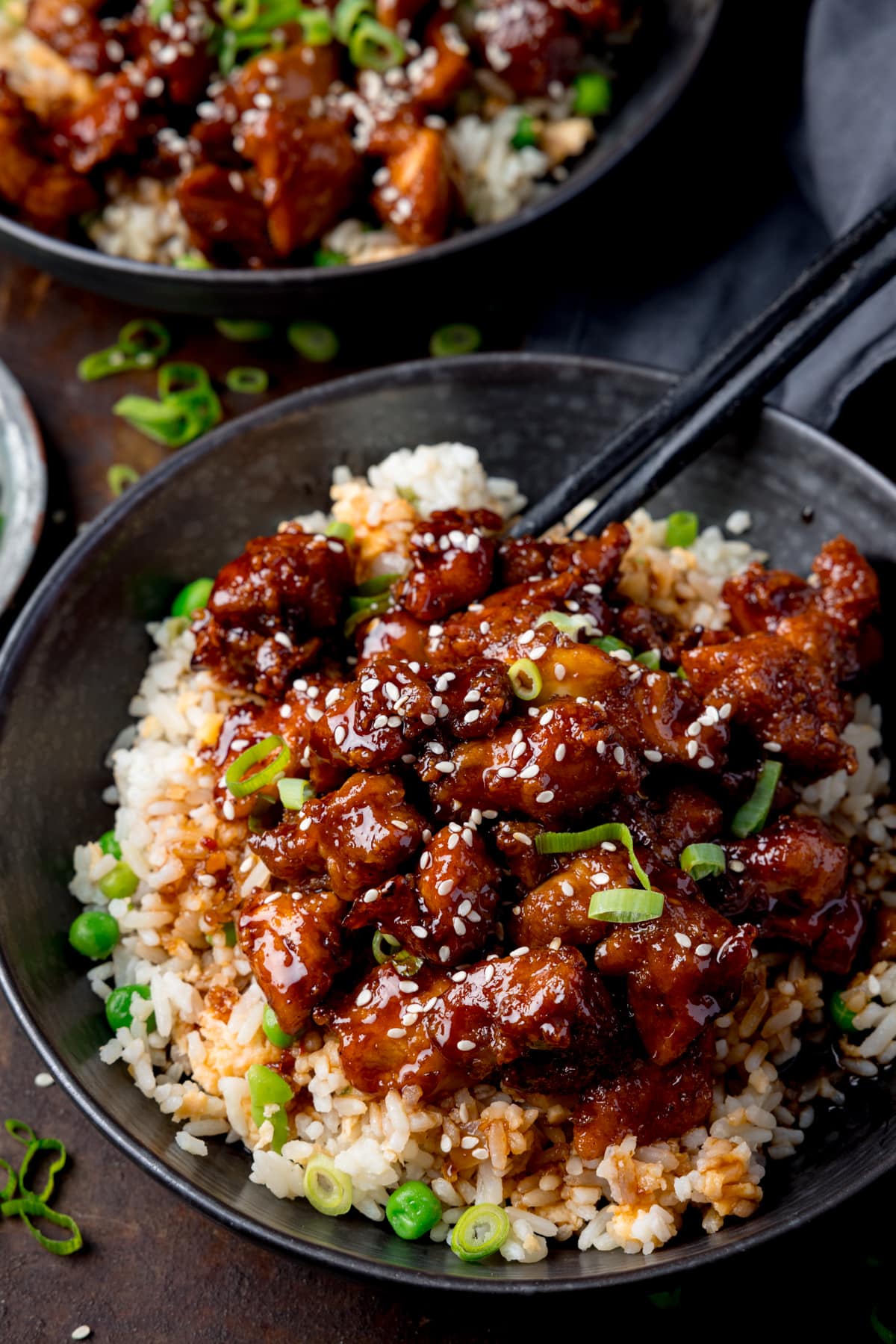Tall image of air fryer sesame chicken served on a bed of egg fried rice in a black bowl. The sesame chicken is topped with sesame seeds and spring onions. There is a pair of black chopsticks sticking out of the bowl. The bowl is on a dark background. There is a further bowl of sesame chicken at the top of the frame.