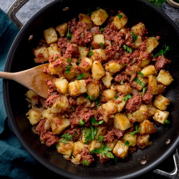 Square overhead image of corned beef hash in a frying pan with a wooden spoon sticking out. There is parsley sprinkled on top.