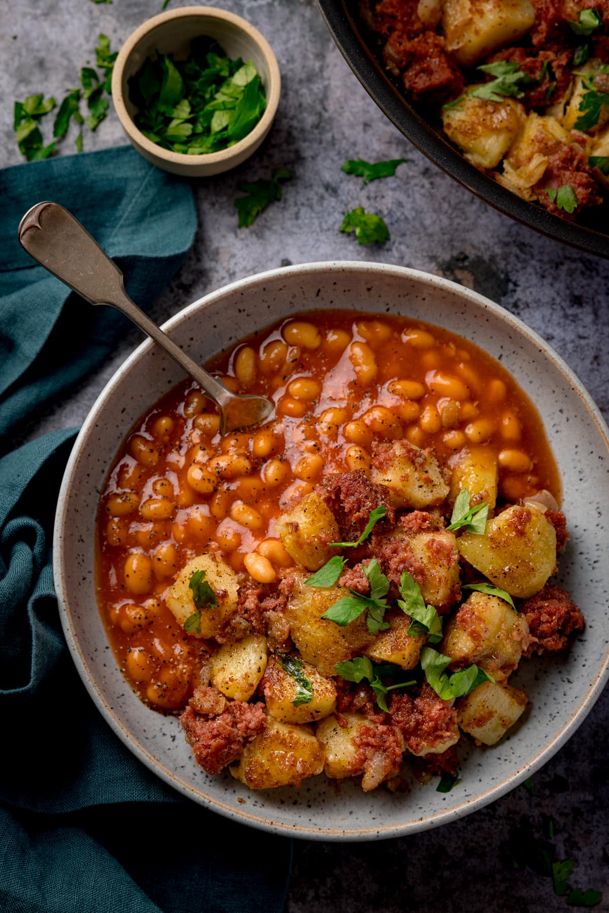 Tall overhead image of corned beef hash with beans in a light bowl with parsley on top. There is a fork sticking out of the beans. The bowl is on a grey surface, next to a green napkin and a small bowl of chopped parsley. There is a frying pan with corned beef hash just in in shot at the top of the image.