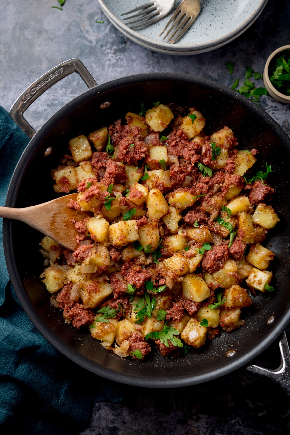 Tall overhead image of corned beef hash in a frying pan with a wooden spoon sticking out. There is parsley sprinkled on top. The pan in on a grey surface next to a dark green napkin. There is a light bowl, a couple of forks and a little bowl with chopped parsley also in shot at the top of the image.
