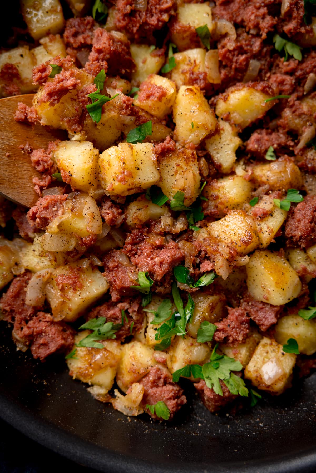 Tall, closeup, overhead image of corned beef hash in a frying pan with a wooden spoon sticking out. There is parsley sprinkled on top.