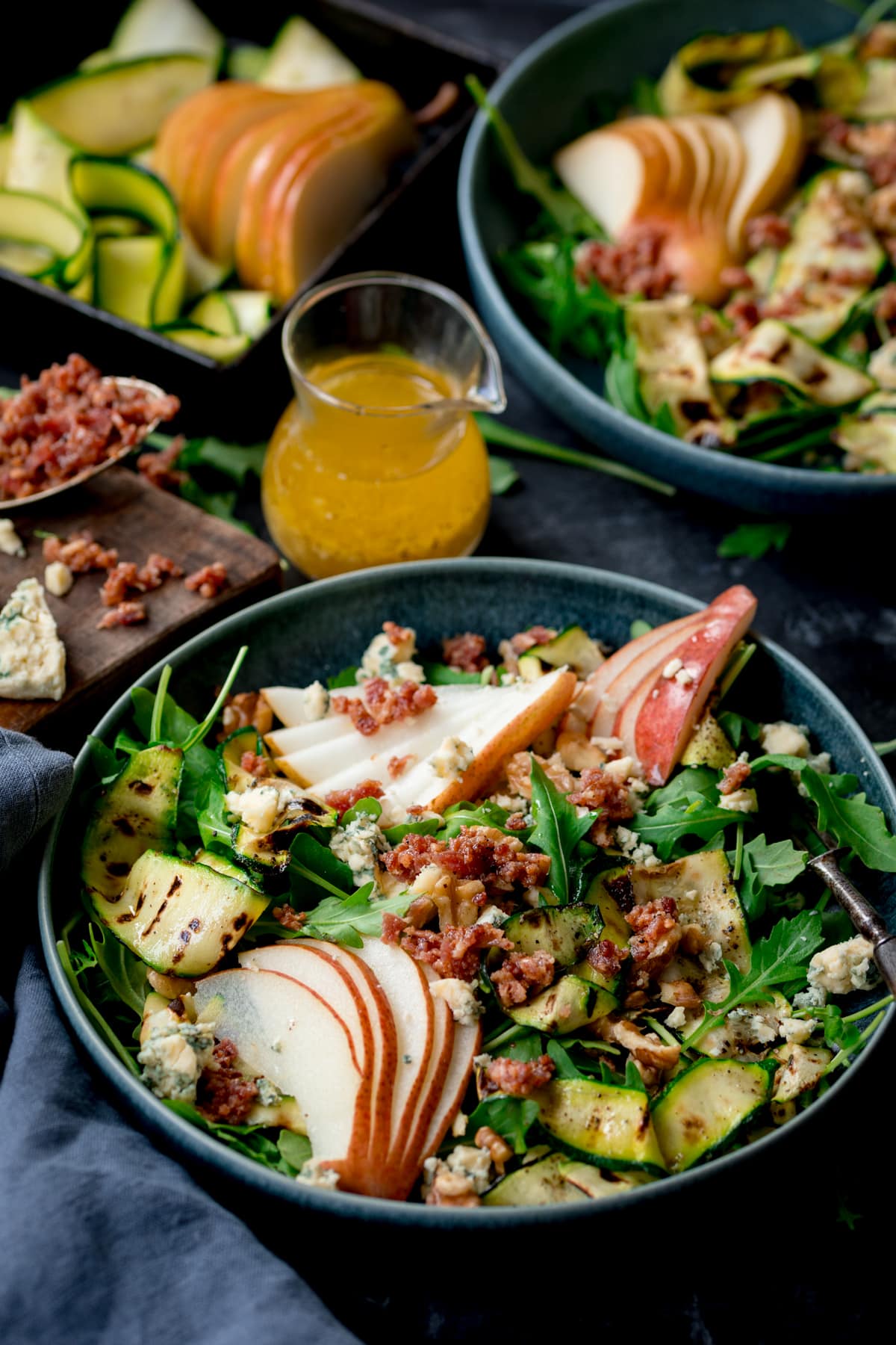 Close up picture of a blue bowl with a grilled courgette salad with sliced pear, crumbled stilton and bacon bits.