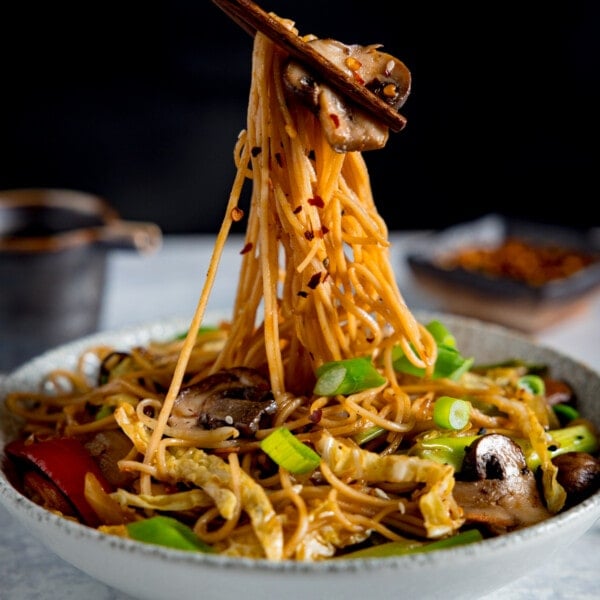 A close-up image of mushroom chow mein in a grey bowl on a grey surface. The mushroom chow mein is topped with spring onions, sesame seeds and chilli flakes. A portion of the mushroom chow mein noodles are being lifted from the bowl by a pair of brown chopsticks. In the background is a small, black jug of chow mein sauce on the left of the frame and a small dish of chilli flakes on the right of the frame. Set on a black background.