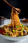 A tall, close-up image of mushroom chow mein in a grey bowl on a grey surface. The mushroom chow mein is topped with spring onions, sesame seeds and chilli flakes. A portion of the mushroom chow mein noodles are being lifted from the bowl by a pair of brown chopsticks. In the background is a small, black jug of chow mein sauce on the left of the frame and a small dish of chilli flakes on the right of the frame. Set on a black background.