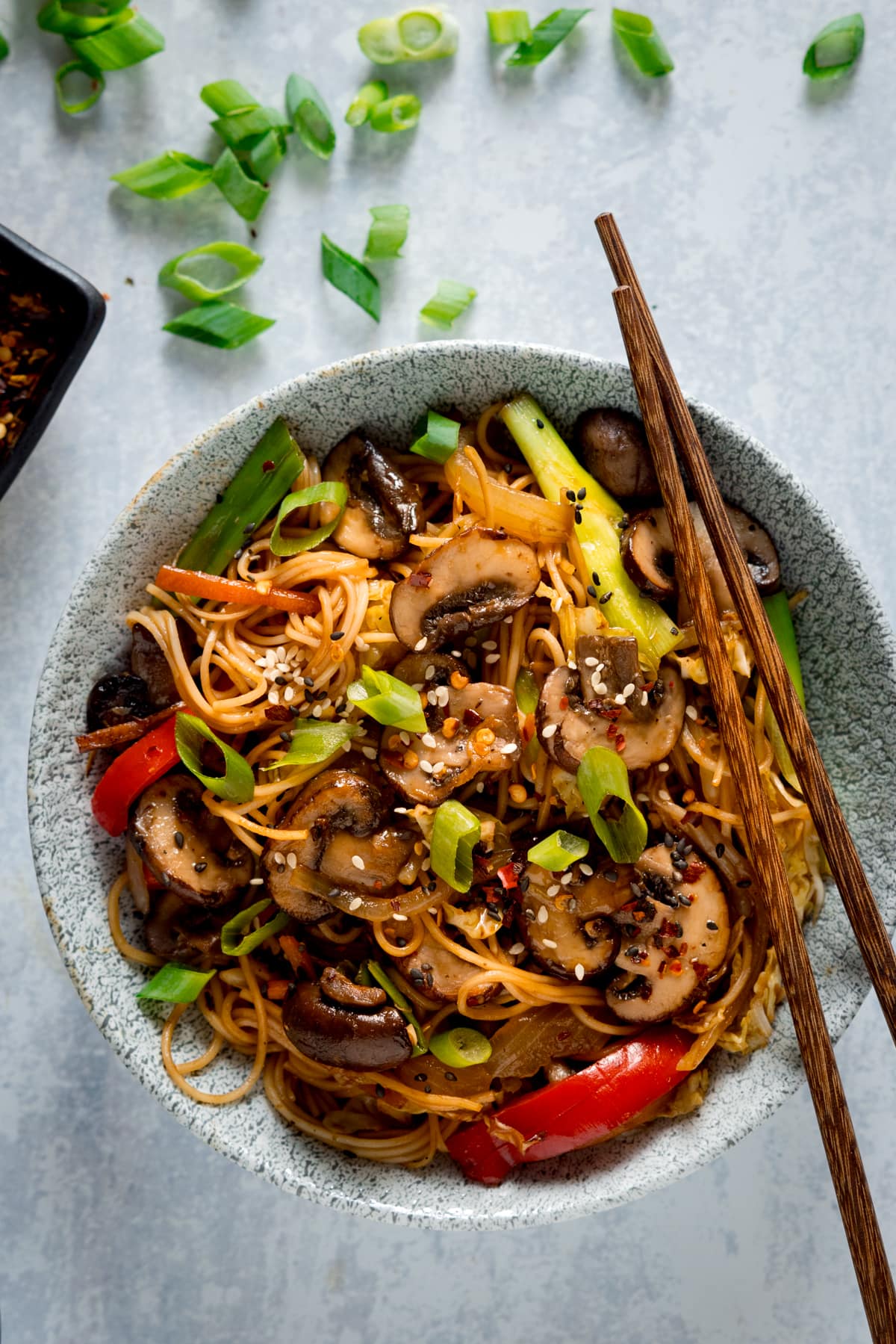 A tall overhead image of mushroom chow mein in a grey bowl on a grey surface. The mushroom chow mein is topped with spring onions, sesame seeds and chilli flakes. There is a pair of brown chopsticks resting on the side of the bowl at an angle. In the background is a small dish of chilli flakes on the left of the frame. There are chopped spring onions scattered around.