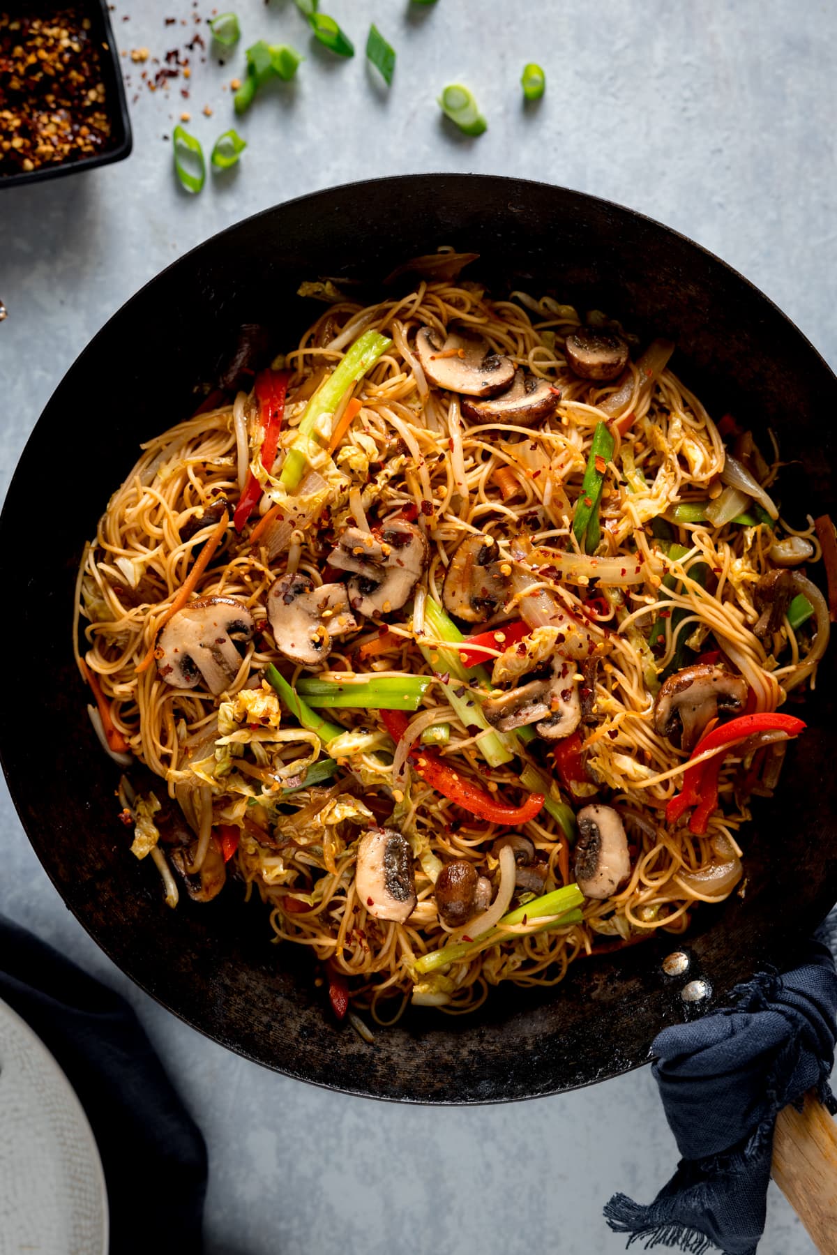 A tall overhead image of mushroom chow mein in a black wok on a grey surface. In the background is a small dish of chilli flakes on the top left of the frame. There are chopped spring onions scattered around. On the bottom left of the frame, there is a grey bowl.