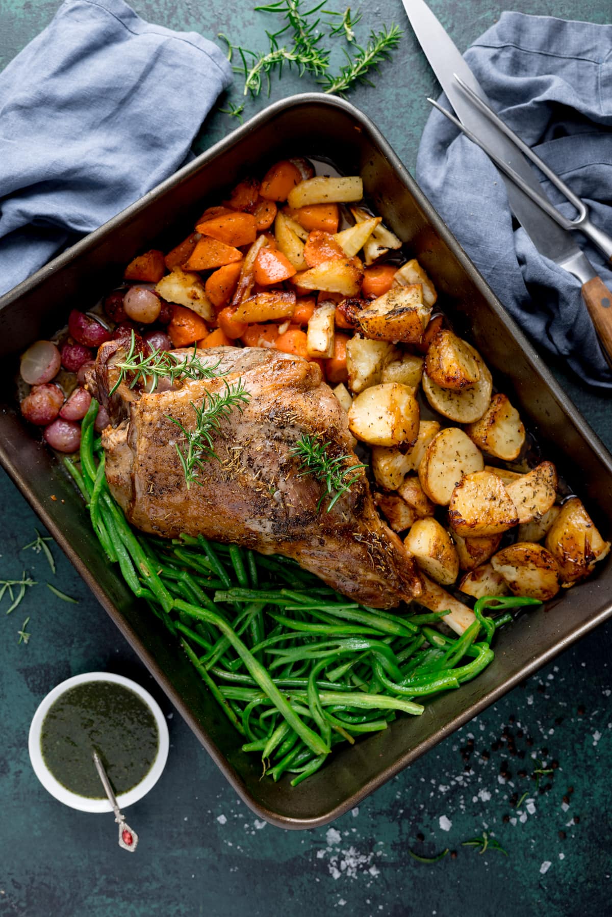 A large roasting pan with a roast leg of lamb with veggies and potatoes in there too garnished with a couple of sprigs of fresh rosemary. The tin is sat on a blue/green board with a bowl of mint sauce in the background.