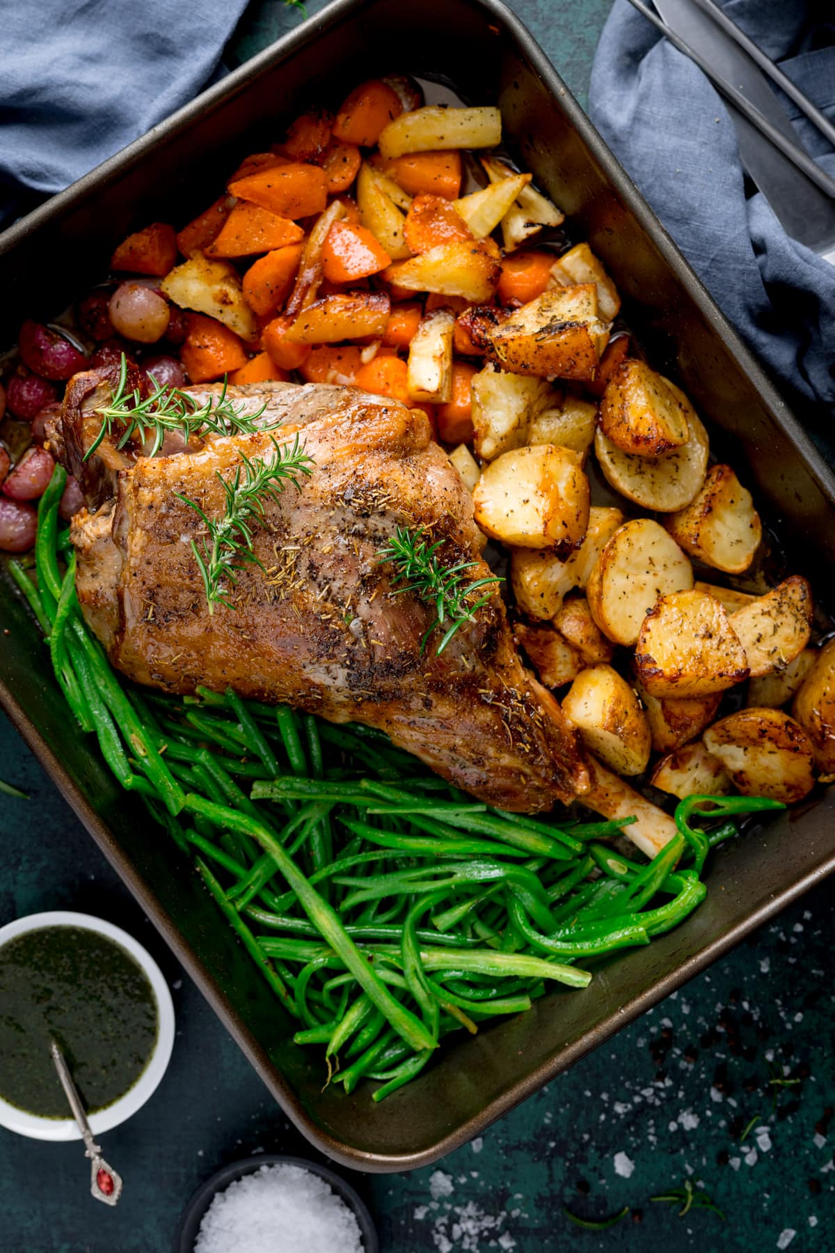 A large roasting pan with a roast leg of lamb with veggies and potatoes in there too garnished with a couple of sprigs of fresh rosemary.