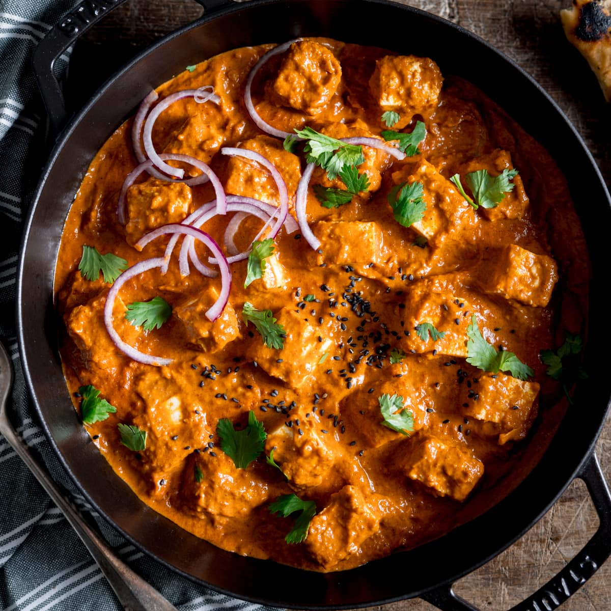 Overhead picture of a paneer curry in a black cast iron pan all sprinkled with some sliced red onion, some fresh coriander and some nigella seeds.