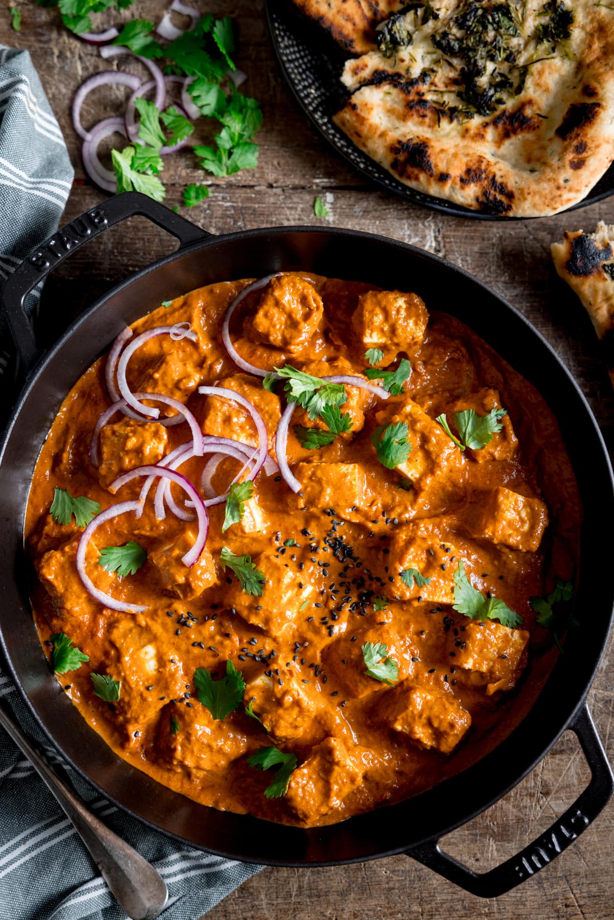 Picture of a paneer curry in black cast iron pan on a wooden board with some garlic naan bread in the background.