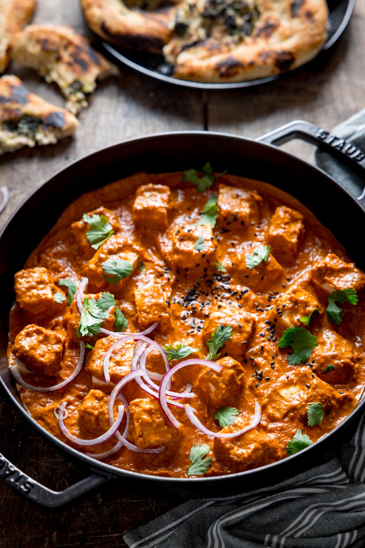 Close up picture oif a creamy paneer curry in a black cast iron pan with some naan bread in the background out of focus.