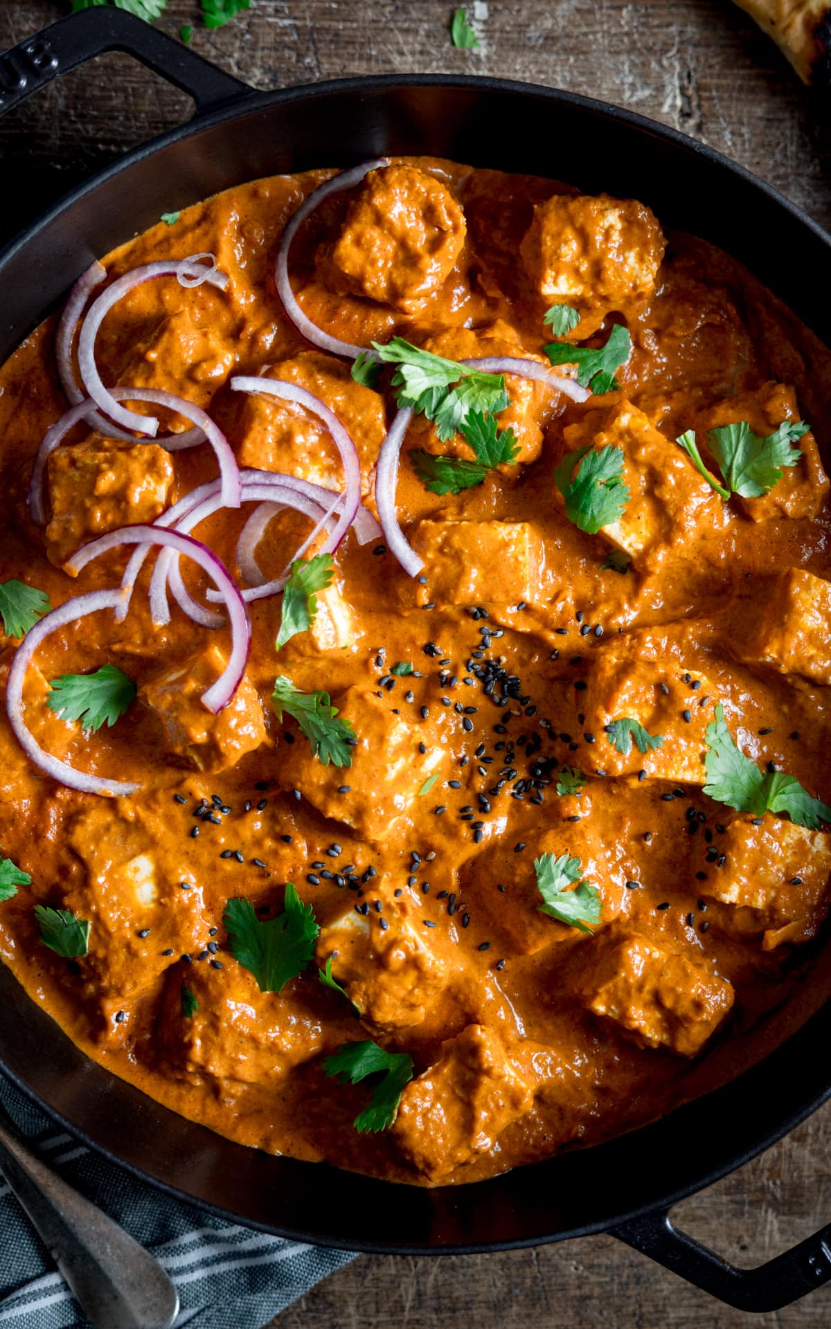 Close up view of a creamy paneer curry in a large skillet with some red onion, coriander and nigella seeds sprinkled on top.