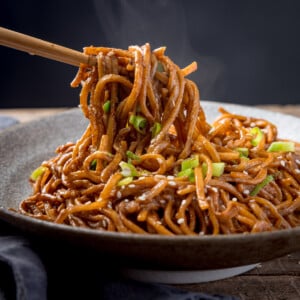 Square image of sesame noodles in a bowl, topped with spring onions and sesame seeds against a dark background. The noodles are being lifted with a pair of wooden chopsticks.