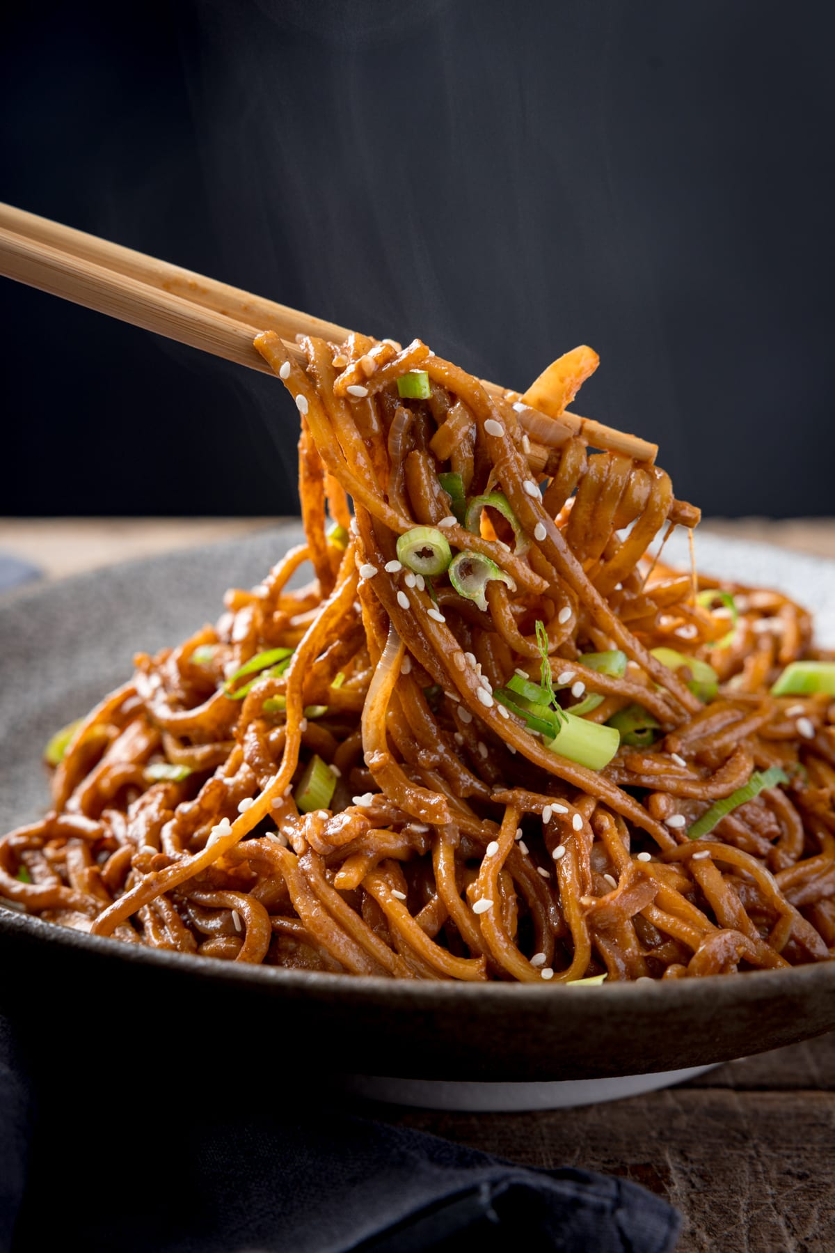 Tall image of sesame noodles in a bowl, topped with spring onions and sesame seeds against a dark background, on top of a wooden surface. The noodles are being lifted with a pair of wooden chopsticks.