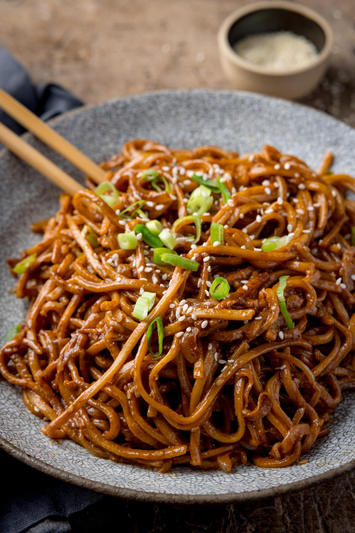 Sesame noodles in a bowl, topped with spring onions and sesame seeds. The bowl is on a wooden table. There is a pair of wooden chopsticks sticking out of the bowl and a little pot of sesame seeds at the top of the image.