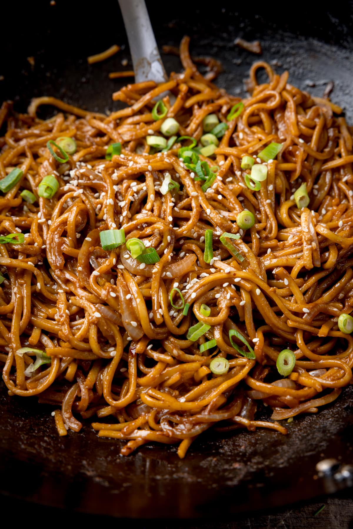 Close up image of sesame noodles topped with spring onions and sesame seeds in a dark wok. The handle of a spatula can be seen sticking out of the noodles.