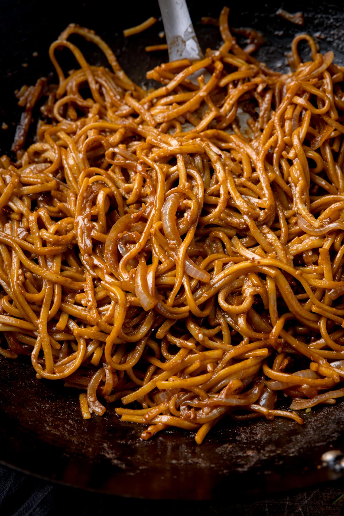 Close up image of sesame noodles in a dark wok. The handle of a spatula can be seen sticking out of the noodles.