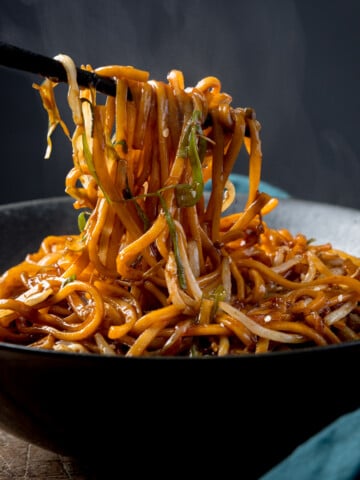 Square side-on image of stir-fried noodles with beansprouts in a black bowl, with sesame seeds sprinkled on top. Some of the noodles are being lifted from the bowl with a set of black wooden chopsticks. There is steam rising from the bowl. The bowl is on a wooden table next to a teal napkin, against a dark background.