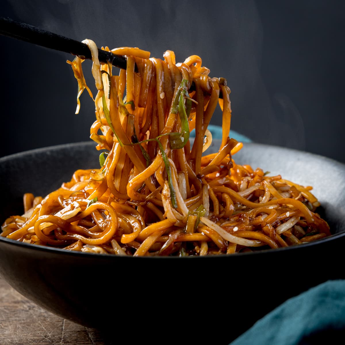 Square side-on image of stir-fried noodles with beansprouts in a black bowl, with sesame seeds sprinkled on top. Some of the noodles are being lifted from the bowl with a set of black wooden chopsticks. There is steam rising from the bowl. The bowl is on a wooden table next to a teal napkin, against a dark background.