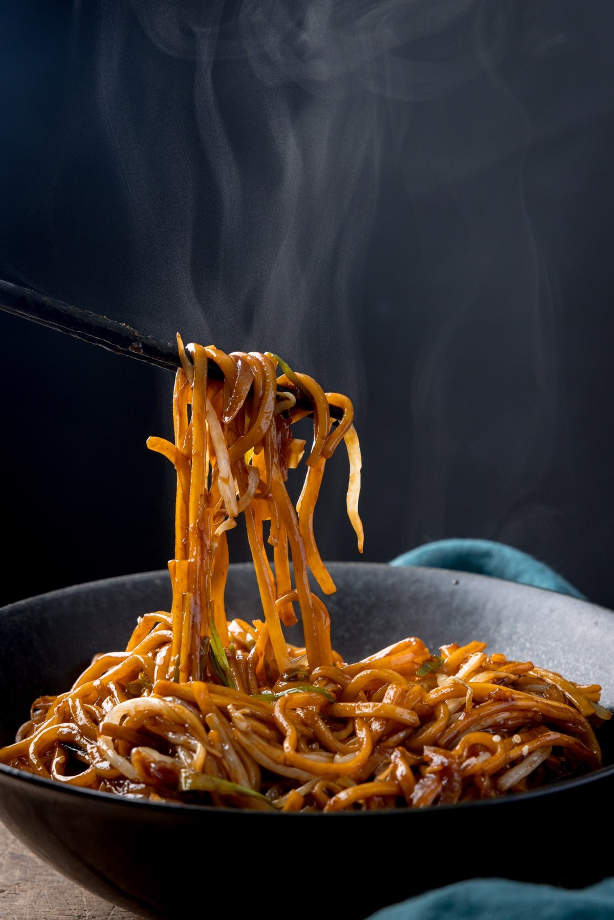 Tall side-on image of stir-fried noodles with beansprouts in a black bowl, with sesame seeds sprinkled on top. Some of the noodles are being lifted from the bowl with a set of black wooden chopsticks. There is steam rising from the bowl. The bowl is on a wooden table next to a teal napkin, against a dark background.