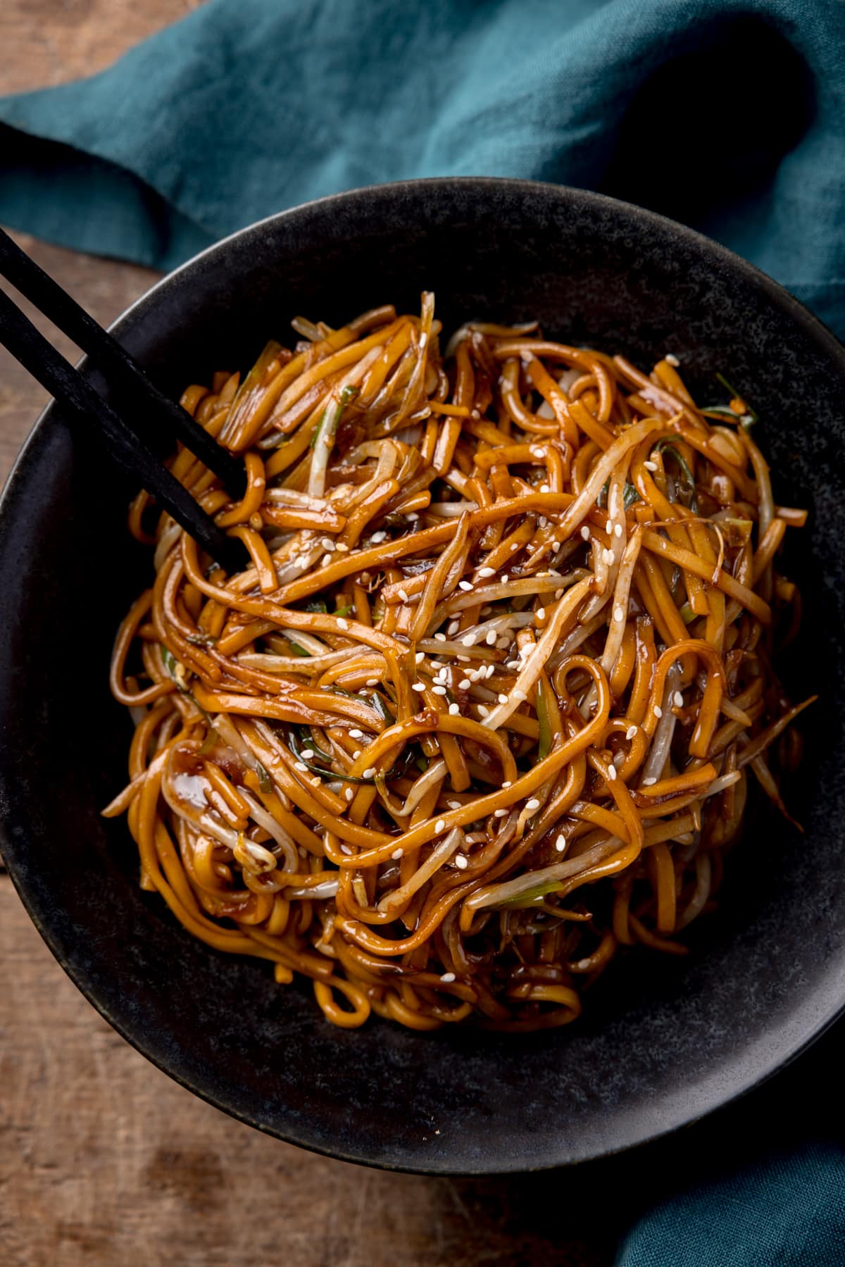 Tall overhead image of stir-fried noodles with beansprouts in a black bowl, with sesame seeds sprinkled on top. The bowl is on a wooden table next to a teal napkin. There is a pair of black wooden chopsticks sticking out of the bowl.